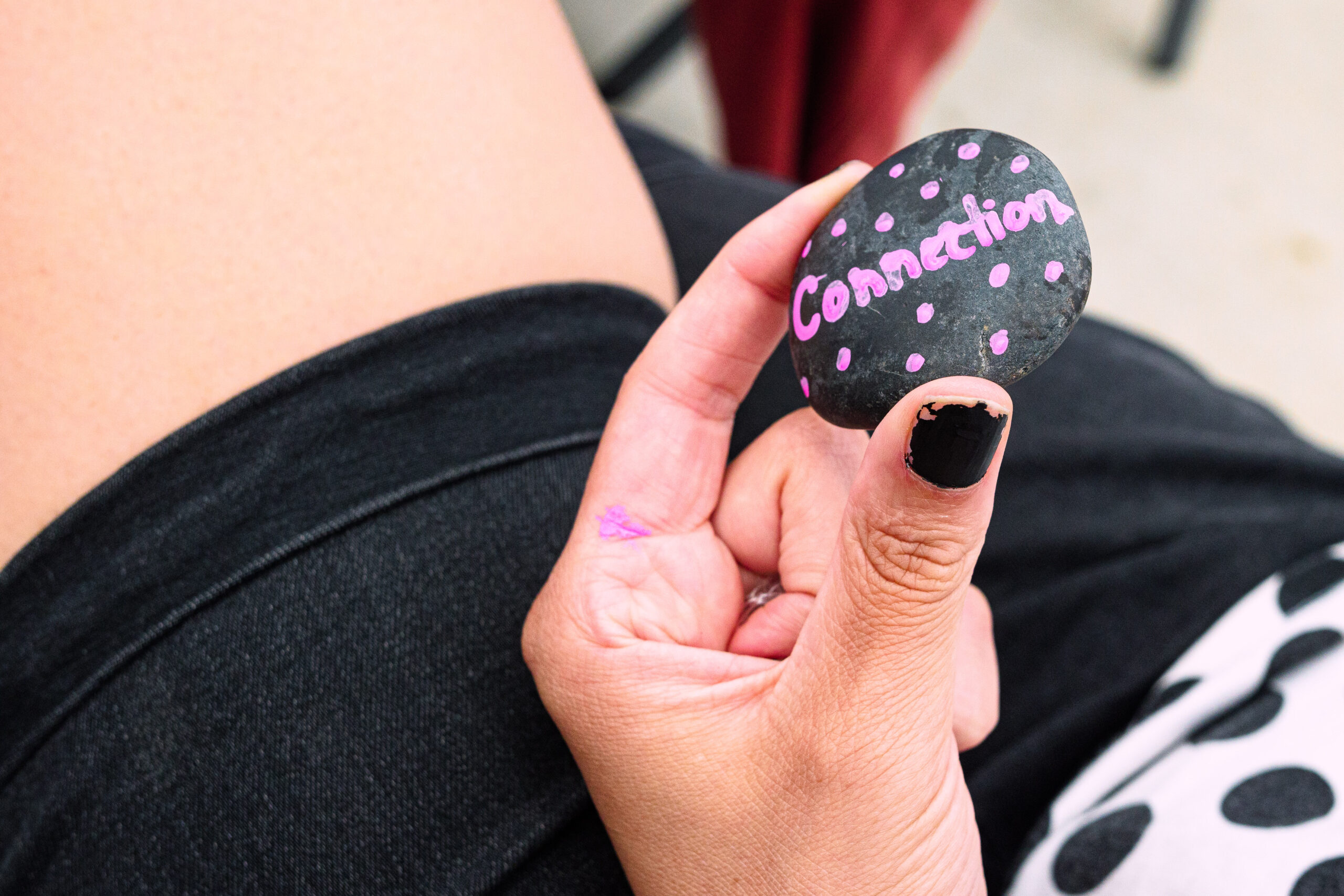 a light skin person in black shorts and a white shirt with black polkadots sits, holding a rock between their thumb and fore finger that has the word "connection" written on it in pink ink. their face is out of view, as the camera is close up and focused on the rock.